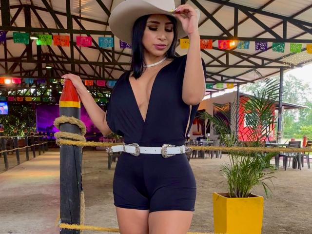 Fransheska Parker indossa una tuta nera con profondo scollo a V, una cintura bianca e un cappello da cowboy, mostrando un seno prosperoso, un sedere formoso e pantaloncini neri, è in piedi vicino a una corda e guarda in basso..