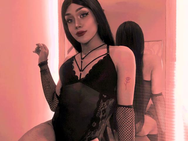 Isabella Veliz, gekleed in zwarte kanten lingerie, met netpanty's en lang zwart haar, met een roos tatoeage op haar rechterarm, staat voor een spiegel. Rode verlichting..