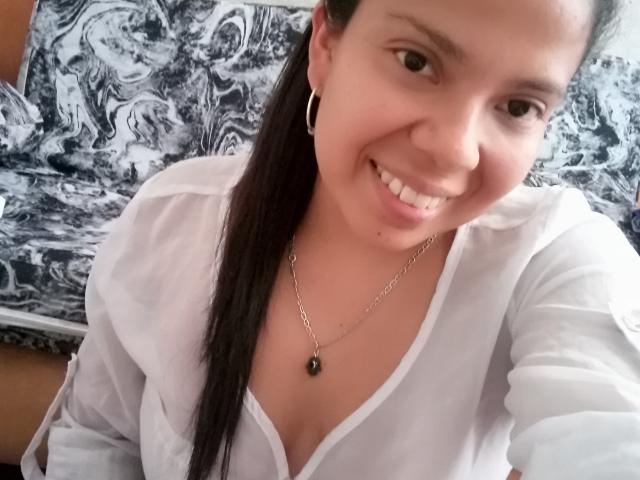 EstrellaBelle ha un seno di media grandezza, sorride indossando una camicia bianca, una collana nera, orecchini a cerchio e una lunga coda di cavallo scura. Sta scattando un selfie, mostrando il suo viso carino e i suoi denti..