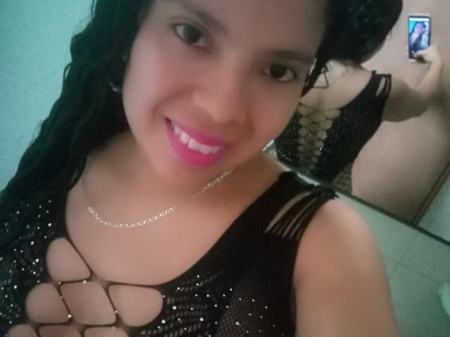 EstrellaBelle con top in rete nera, che mostra i suoi seni attraverso un motivo traforato, sorride in un selfie allo specchio con rossetto rosa, tiene in mano il telefono, capelli neri ondulati, riflesso nello specchio..