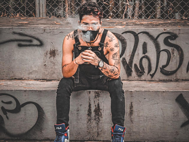 SteveArt, tatuado, fumando, usa máscara preta, suspensórios e tênis azuis Fila, está sentado em concreto sujo, segurando um telefone, com tatuagens visíveis e braços musculosos, em um ambiente urbano degradado..