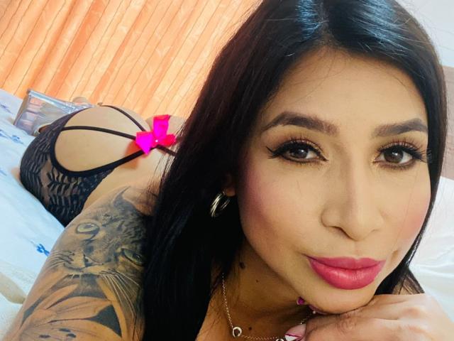 Fransheska Parker, con i capelli scuri, il rossetto rosa e il braccio tatuato, sdraiata sul letto, indossa biancheria intima di pizzo nera, ha un adesivo a forma di cuore rosa sui glutei, guarda la fotocamera con un'espressione accigliata..