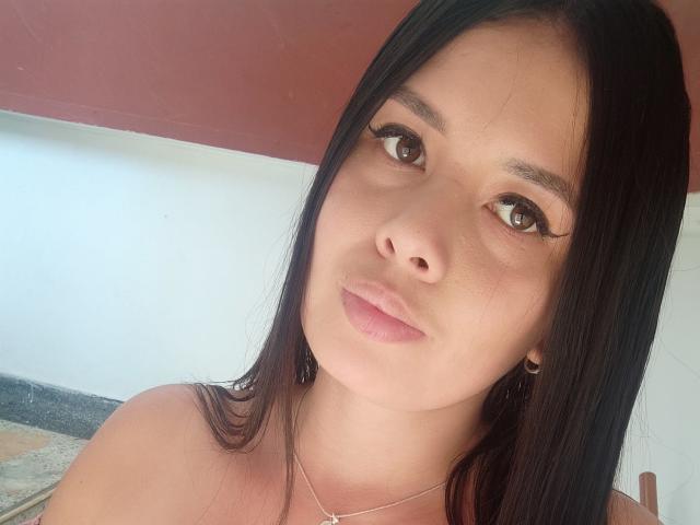 Het gezicht van AshleyKatelyn, met lang zwart haar, bruine ogen en roze lippen, draagt een zilveren ketting en kijkt met een lichte glimlach naar de camera. Haar borsten zijn niet zichtbaar..
