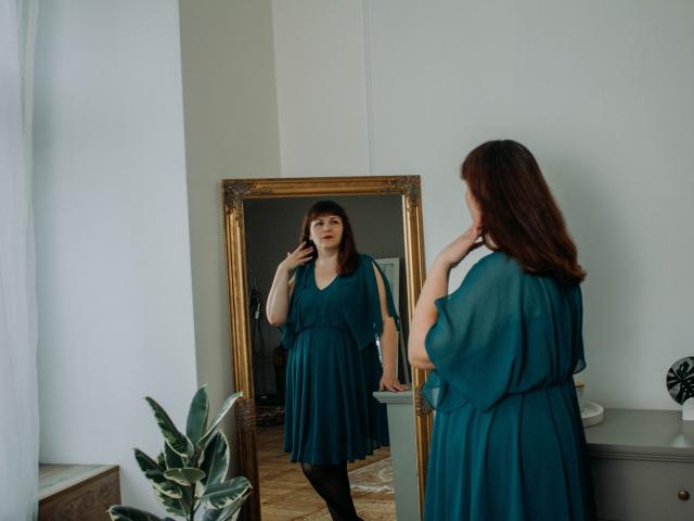 AliceInks stands in front of a mirror, wearing a teal dress, looking at her reflection. She has brown hair, and her tits are visible in the mirror.