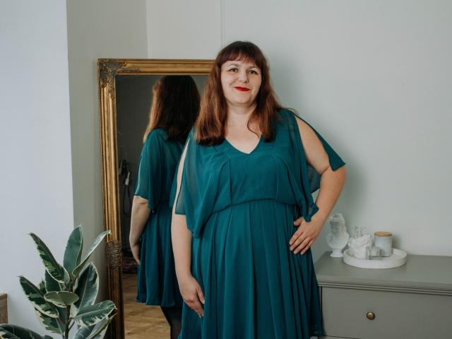 AliceInks, a chubby woman with red lipstick, wearing a teal dress, standing in front of a mirror, her hand on her hip, showing her ass reflected in the mirror.