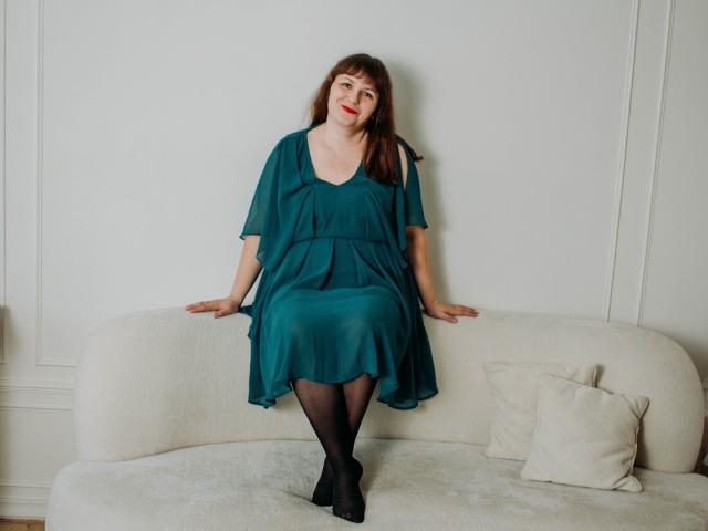 AliceInks with red lipstick, brown hair, teal dress, black stockings, sitting on white couch, hands on seat, looking at camera, subtle smile, casual, cute, attractive.