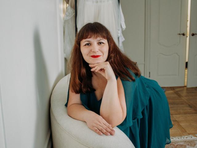 AliceInks with red lipstick, brown hair, and teal dress, resting her chin on her hand, smiling, sitting on a white curved chair, wooden floor, white wall.