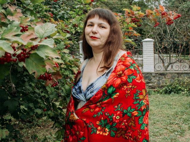 AliceInks has medium tits, wearing a red floral shawl over a low-cut blue shirt, standing in a green garden with red berries and leaves. She's smiling with red lipstick.
