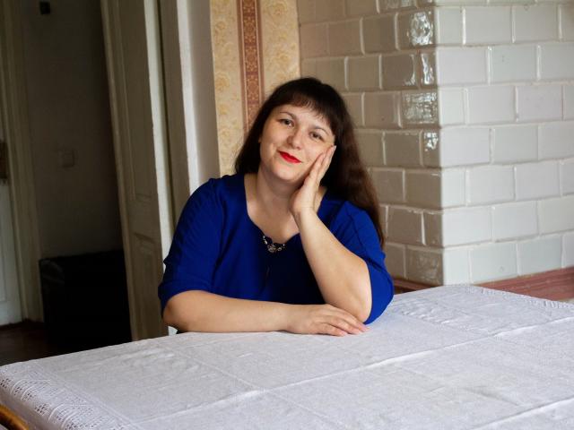 AliceInks with red lipstick, blue shirt, and dark hair, resting her cheek on her hand, sitting at a white table, looking slightly to the side, smiling subtly.