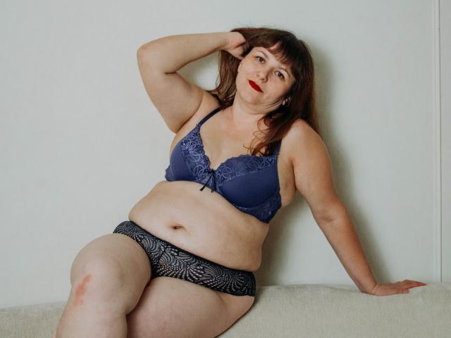 AliceInks, plus-size with pale skin, dark brown hair, red lipstick, wearing blue lace bra, black patterned panties, sitting on white couch, hand in hair, looking at camera.