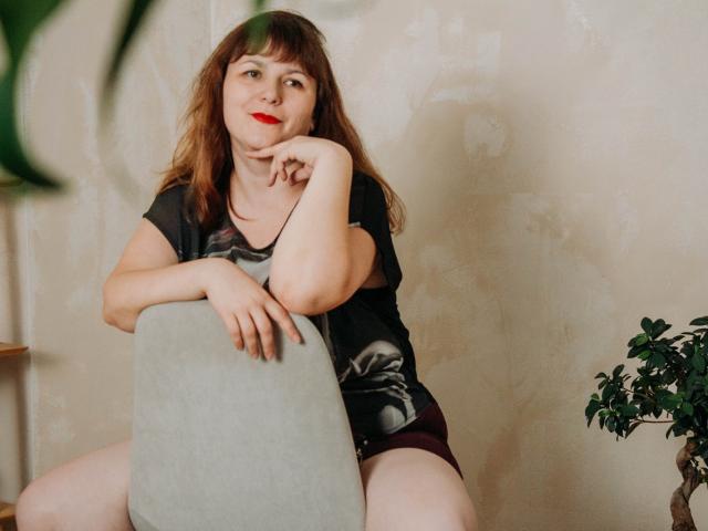 AliceInks with red lipstick, sitting on a gray chair, black t-shirt, and maroon shorts, resting her chin on her hand, brown wavy hair, slightly smiling, with green plant.