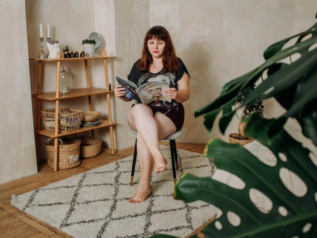 AliceInks, with red hair and tits covered by a black shirt, sits barefoot on a chair reading a magazine, legs crossed, in a wooden room with a plant.
