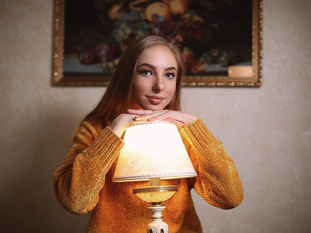 SafiraFuracao mit blonden Haaren, blauen Augen und einem süßen Lächeln, hält eine angezündete Lampe, trägt einen orangefarbenen Strickpullover, ihre Hände ruhen auf der Lampe. Ihre Brüste sind klein..