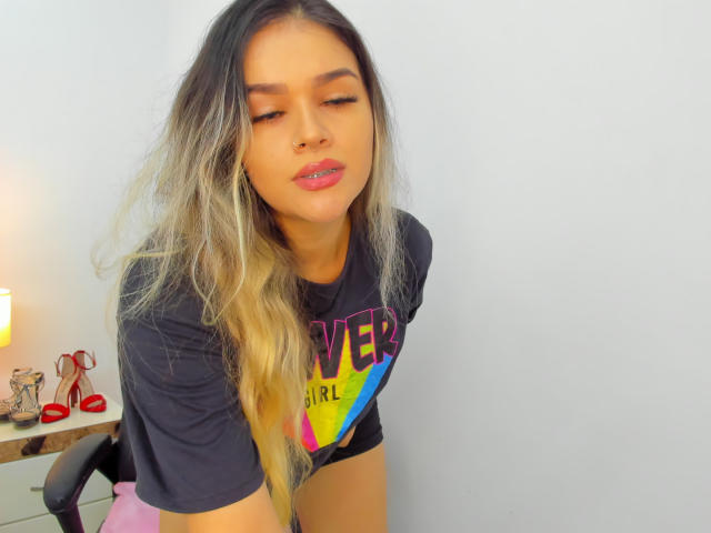 ValerieStone with long, ombre hair, closed eyes, and pink lipstick, wearing a black "LOVER GIRL" shirt, leaning forward, showing her small, round ass, in a brightly lit room.