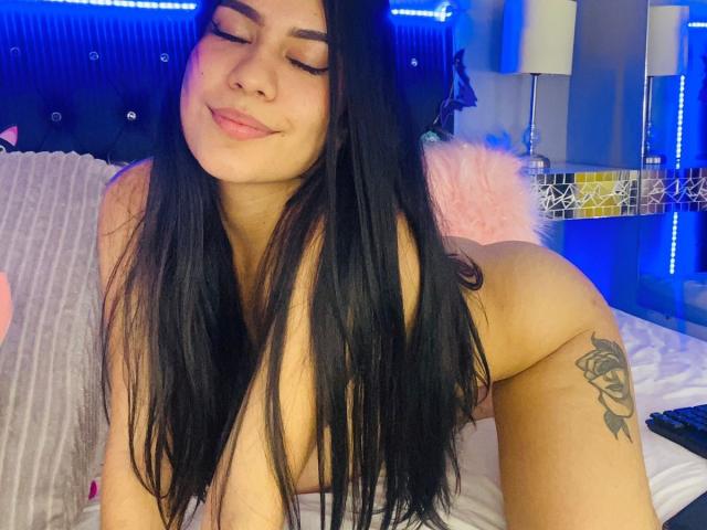 GabbySumer met lang zwart haar, ogen gesloten, zacht lachend, naakt, met grote borsten, roos tatoeage op de rechter dij, op een bed met blauwe LED-verlichting op de achtergrond..