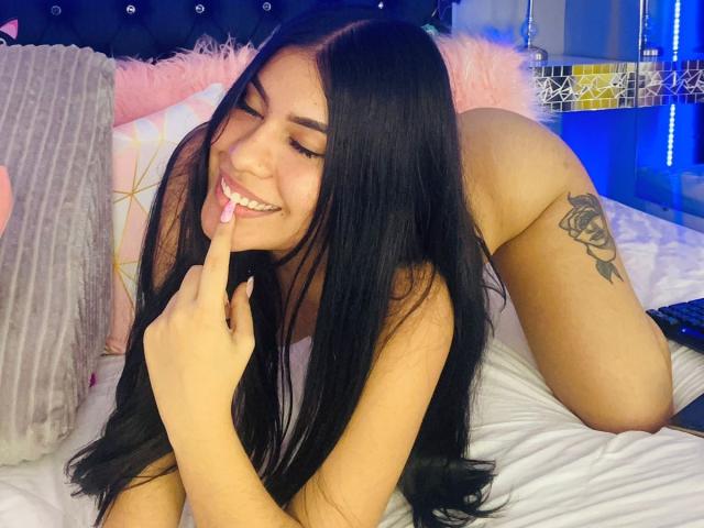 GabbySumer met lang zwart haar, lachend, met een vinger in haar mond, op bed, naakt, met een tatoeage op de rechter dij, blauw licht in de slaapkamer, wit bed, roze kussen, grijze deken, lachend..