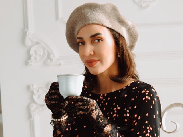 SimoniGrace, com um chapéu cinza, luvas de renda preta e um vestido floral, segurando uma xícara de chá branca, exibindo discretamente suas nádegas com um sorriso sutil..