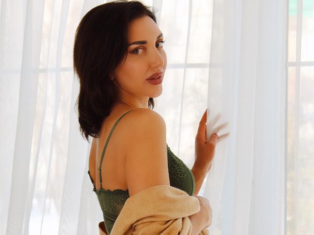 SimoniGrace con capelli castano scuro, reggiseno di pizzo verde e cappotto color sabbia, che tiene una tenda bianca, guarda oltre la spalla, luce soffusa, leggero sorriso, schiena scoperta, carina, sexy..