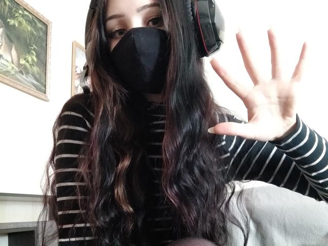 MeliMuller, con cabello largo y negro, vestida con un top corto blanco, una falda escocesa roja y calcetines blancos hasta las rodillas, sentada en una cama, sosteniendo un teclado negro, mostrando su trasero..
