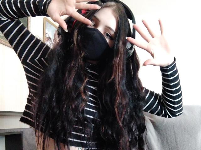 KirishikiRetta, con el pelo largo, ondulado y oscuro, vistiendo una máscara negra y una camisa a rayas, auriculares negros, levantando una mano, mostrando un rostro lindo, seguro y sexy..