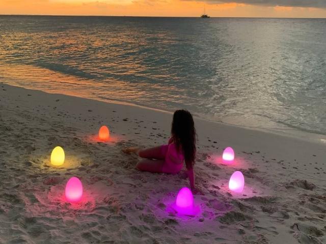 BellaFlower con un bikini rosa, sentada en una playa arenosa con orbes moradas y rosas brillantes a su alrededor, mirando un atardecer sobre el océano. Su trasero es ligeramente visible..
