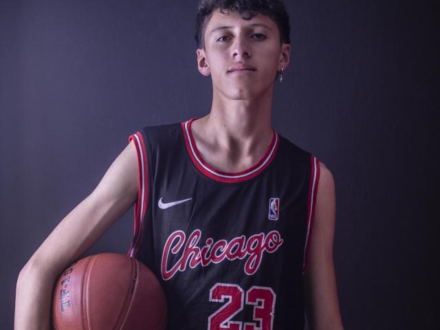 Carlos Rubiio, avec des cheveux courts et bouclés bruns, portant un maillot de basket noir de Chicago, numéro 23, tenant un ballon de basket sous son bras droit, fond sombre..