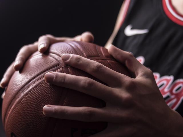 CarlosRubiio's hands gripping a brown basketball, showing fingers on the textured leather, wearing a black Nike basketball jersey with red and white accents.
