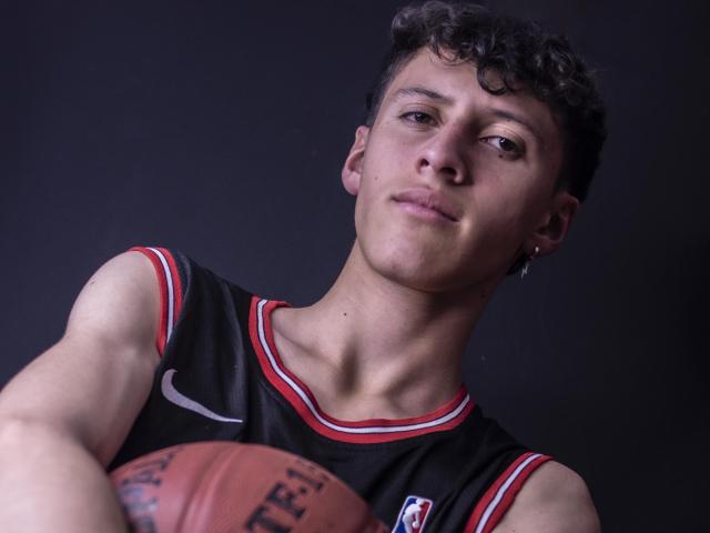 CarlosRubiio, holding a basketball in front of him, wearing a black sleeveless jersey with red trim, looking down, slightly smiling, short curly brown hair, dark background.