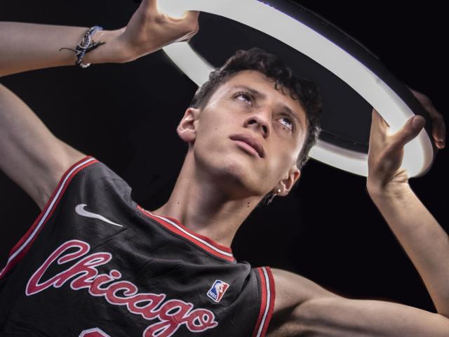 Carlos Rubiio, verschwitzt, mit lockigem, braunem Haar, trägt ein Chicago Basketball-Trikot, hebt einen Korb über seinem Kopf, blickt nach oben, dunkler Hintergrund, schwarzer Ärmel, silbernes Armband..