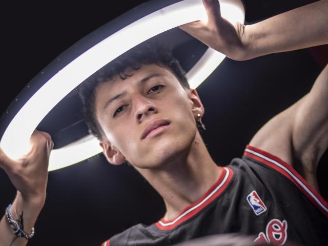 Carlos Rubiio houdt een ringlamp boven zijn hoofd, kijkt met een lichte glimlach naar de camera en draagt een donkergrijze basketbalshirt met rode en witte accenten..