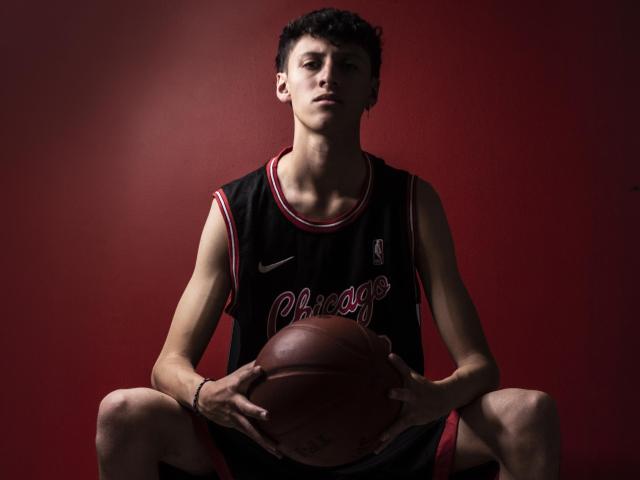 Carlos Rubiio mit lockigem, schwarzem Haar, der ein schwarzes Basketballtrikot trägt, einen Basketball zwischen seinen Knien hält, in die Hocke gegangen ist, ernst aussieht, roter Hintergrund, Halbnahaufnahme, konzentrierter Gesichtsausdruck..