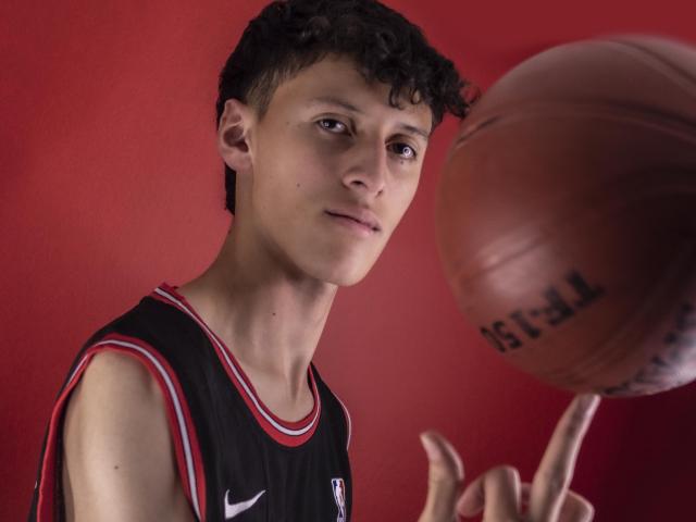 Carlos Rubiio, met krullend zwart haar, draagt een zwart en rood basketbalshirt, houdt een basketbal vast, wijst met zijn rechter wijsvinger ernaar, rode achtergrond, geconcentreerde uitdrukking, medium shot..