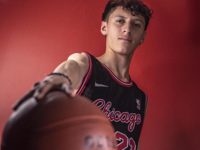 Carlos Rubiio, met krullend bruin haar, gekleed in een zwart Chicago Bulls shirt, die een basketbal vasthoudt in een lage hoek, rode achtergrond, geconcentreerde uitdrukking, rechterarm uitgestrekt naar voren..