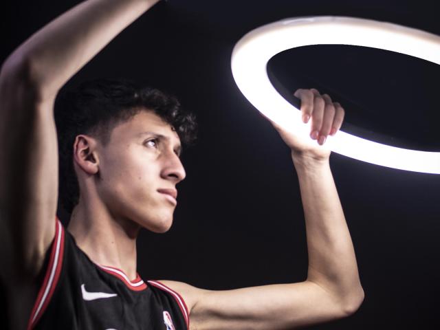 CarlosRubiio with curly black hair, wearing a black Nike basketball jersey, holding a glowing white ring light above his head, looking up with a focused expression.