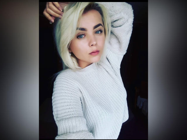 EvaEflii with blonde hair, blue eyes, and a nose ring, wearing a fluffy white sweater, raising her arm, looking at the camera with a slightly parted lip.