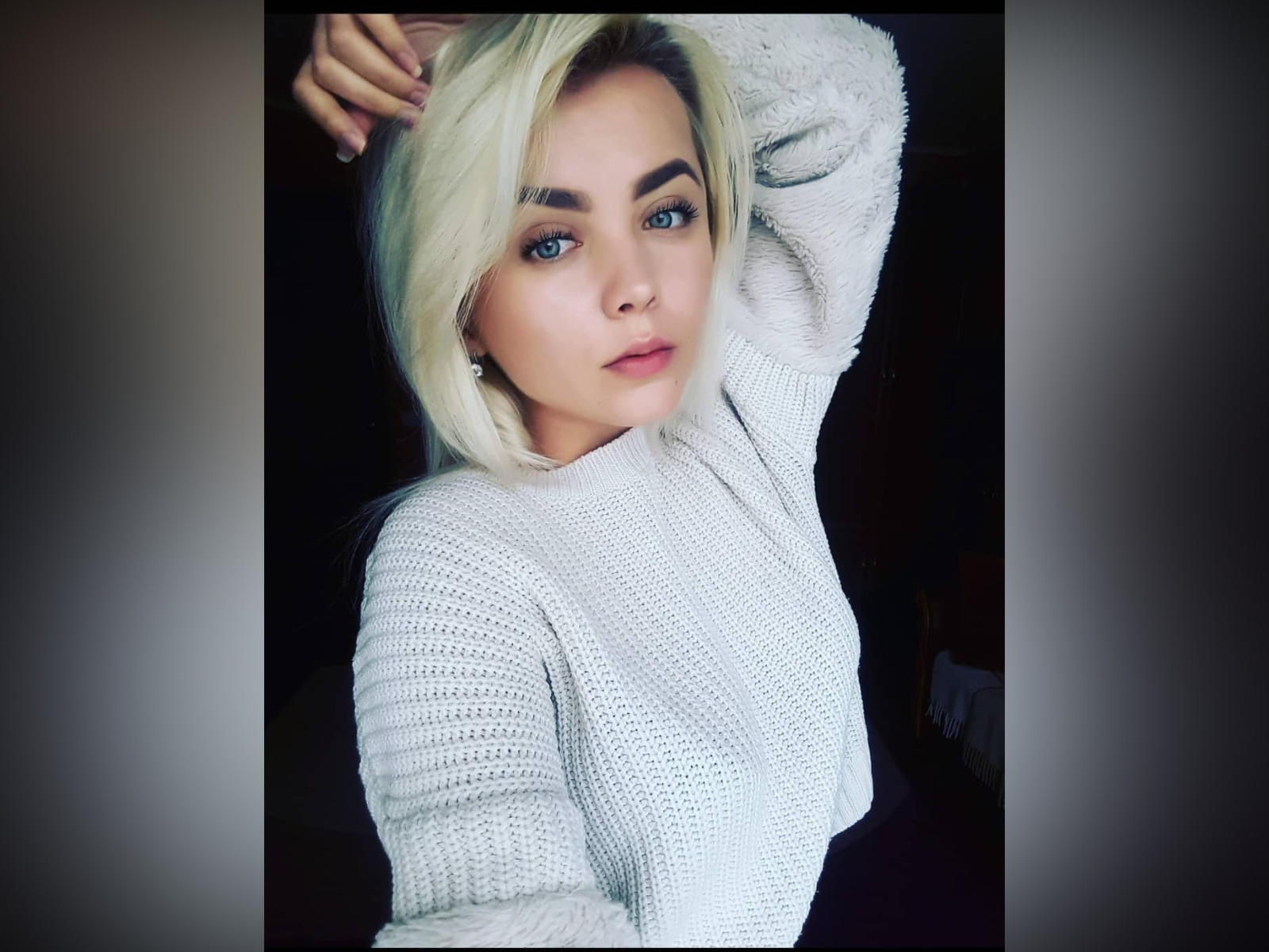 EvaEflii mit blonden Haaren, blauen Augen und einem Nasenring, trägt einen flauschigen weißen Pullover, hebt ihren Arm und blickt mit leicht geöffnetem Mund in die Kamera..