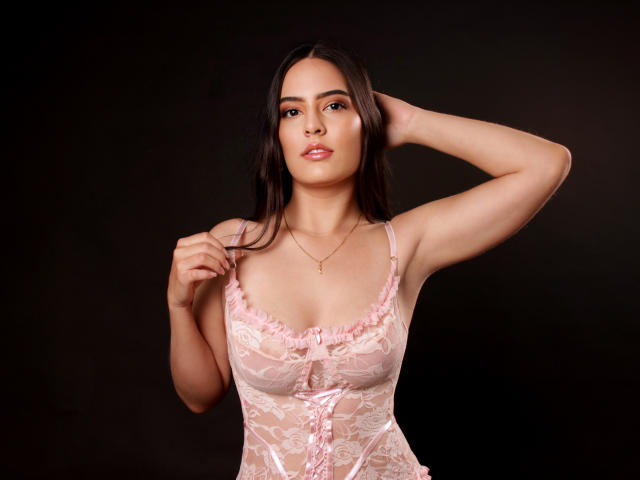 FallonJohnson, con senos de tamaño medio, vestida con lencería de encaje rosa, tiene una mano en el pelo, ojos marrones, cabello largo y oscuro, una ligera sonrisa, fondo negro, collar dorado, luz suave..