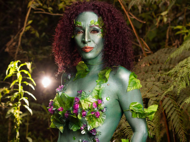 KhazandraX with green-painted skin, curly red hair, and leaf decorations covering her tits, standing in a jungle-like setting, looking at the camera with an orange lipstick smile.