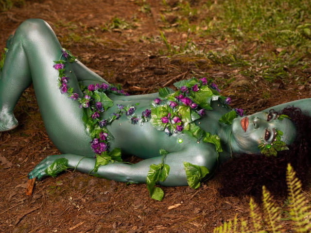 KhazandraX, green-skinned with purple flowers on her tits and pussy, lies on forest floor, arms above head, legs spread, green leaves on body, curly dark hair, forest floor background.