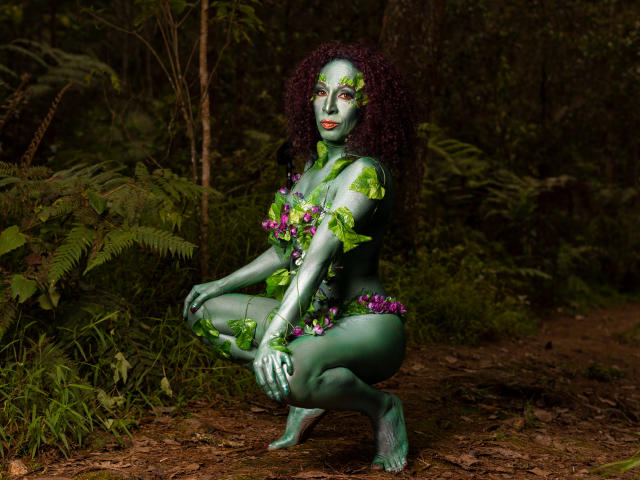 KhazandraX, green-skinned with leaf and flower tattoos, crouches in forest, naked, with curly purple hair, green eyes, and purple flowers covering her tits and pussy.