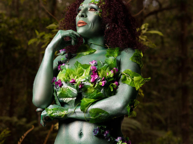 KhazandraX with green-painted skin, curly red hair, leafy vines covering her tits and arms, wearing purple flowers, looking up with orange lipstick in a forest.