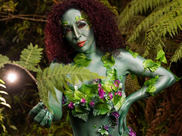 KhazandraX with green-painted skin, curly red hair, and leafy decorations, showing off her boobs and purple flowers, standing in a jungle-like setting with ferns.