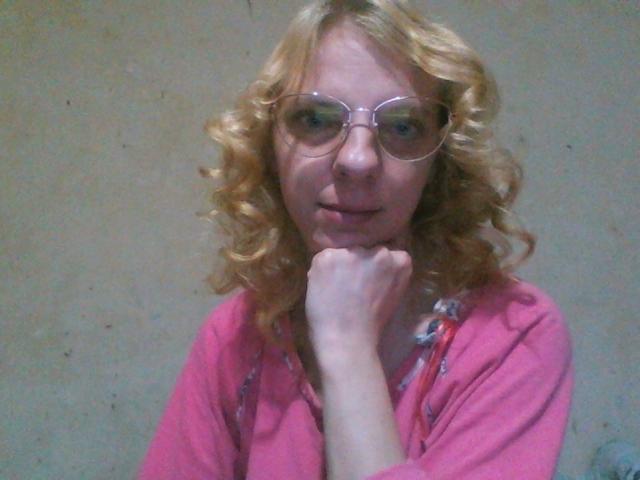 BlondyMarianne, con cabello rubio rizado, vestida con ropa rosa, gafas, y con el mentón apoyado en la mano, luciendo ligeramente cansada. Su camisa rosa tiene un estampado floral..