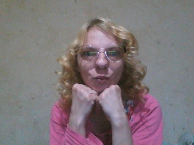 BlondyMarianne, con cabello rubio rizado, una camisa rosa y gafas transparentes, frunciendo los labios, con las manos debajo del mentón, mirando a través de las gafas, mostrando ligeramente sus senos, en una fotografía con iluminación suave..