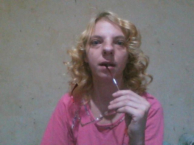 BlondyMarianne, avec ses cheveux blonds bouclés, sa chemise rose et ses lunettes, suce un rouge à lèvres rouge, la bouche légèrement ouverte, et regarde vers le haut. Ses seins sont petits et elle est mignonne..