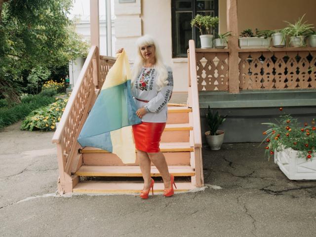 KamaliaSim with fluffy white hair, gray sweater, red leather skirt, red heels, holding colorful blanket, standing on pink steps, house with potted plants, and flowers in background.