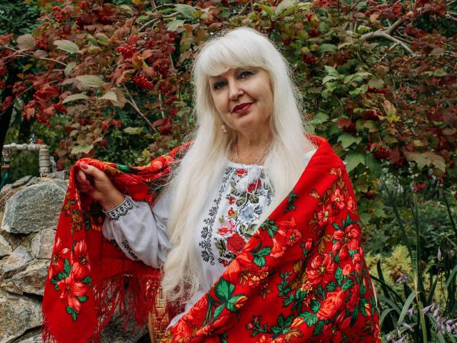 KamaliaSim with long white hair, wearing a red floral shawl, grey embroidered shirt, holding shawl open, smiling, standing by a stone wall, outdoors, autumn foliage.