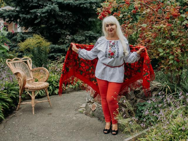 KamaliaSim with long blonde hair, gray shirt with red floral embroidery, red pants, holding red floral blanket, standing on stone path, wicker chair to left, smiling.