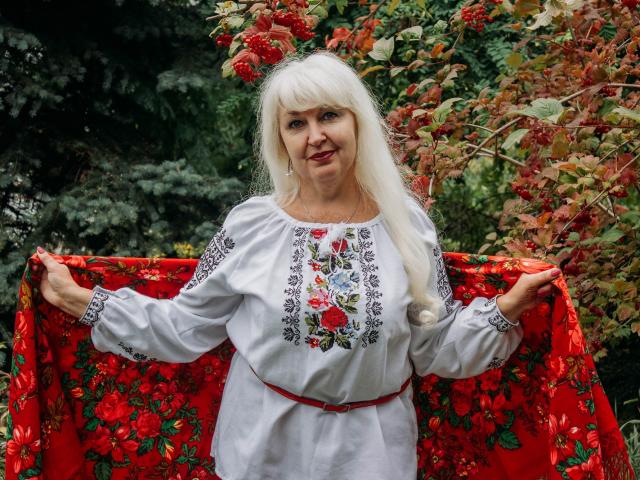 KamaliaSim with long white hair, smiling, wearing a white dress with red and floral embroidery, holding a red floral fabric, standing outdoors, showing tits with dress open.