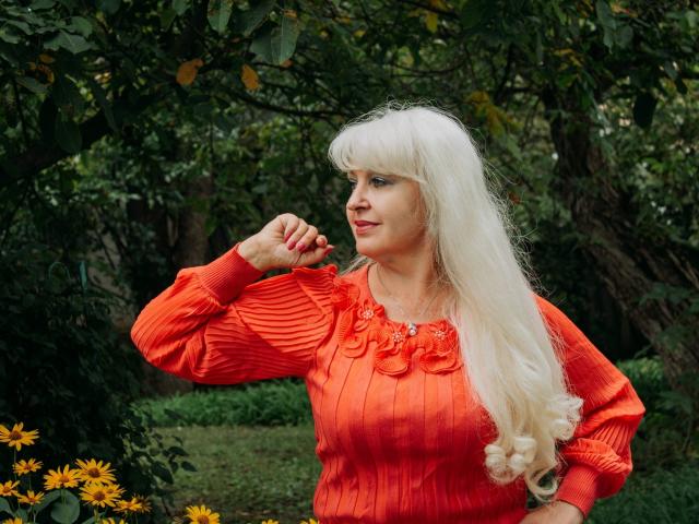 KamaliaSim with long, wavy, white wig, wearing an orange, ribbed, long-sleeve shirt, raising her right hand, standing in a garden with yellow flowers and green foliage.